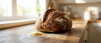 Freshly baked artisan bread loaf with crispy crust resting on wooden kitchen counter near window, ideal for bakery, food, and culinary content.