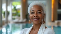 Confident mature African American woman with short white hair and gold hoop earrings smiling warmly at spa resort, wearing white bathrobe against blurred pool background.