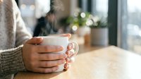 Woman hands holding steaming hot coffee mug in cozy cafe atmosphere with warm bokeh lights creating peaceful morning moment for relaxation and comfort.