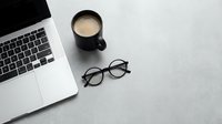 Minimalist workspace flat lay with laptop, black coffee mug, and round glasses on a gray concrete surface.