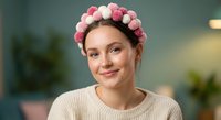 Young Caucasian woman wearing pink flower crown headband smiling warmly in cozy indoor setting with soft lighting and plants in background.