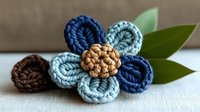 Handcrafted crochet flower in blue and brown tones with green leaves, close-up view on white surface, showing detailed textile texture and artisanal design.