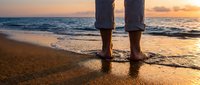 Barefoot person standing on sandy beach at golden sunset, waves gently washing over feet, peaceful seaside moment evoking freedom and relaxation.