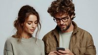 Young couple looking at smartphone together, woman with loose updo hairstyle and man with glasses and beard wearing casual earth-tone clothing against neutral background.