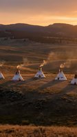 Traditional teepees in golden prairie landscape at sunset with rolling hills and warm orange sky creating peaceful camping scene for outdoor adventure concepts.