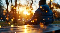Person in winter jacket sitting alone on bench at sunset with golden bokeh lights and lens flares creating atmospheric mood for contemplation and solitude.
