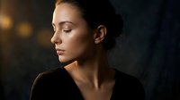 Young woman in profile with closed eyes and serene expression illuminated by warm golden light against dark background for wellness and meditation content.