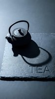 Cast iron teapot on dark slate board with TEA inscription, dramatic shadow, moody dark aesthetic for tea ceremony, menu, or wellness branding.