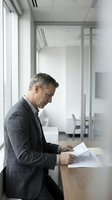 Middle-aged Caucasian male businessman in dark suit reviewing financial documents at wooden desk in modern office with glass partitions and natural light from windows.