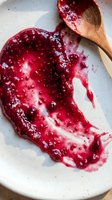 Homemade berry jam spread on white ceramic plate with wooden spoon, showing vibrant red fruit preserve for cooking and baking projects.