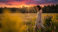 Woman in white dress standing in wildflower meadow at golden hour sunset, surrounded by blooming flowers and forest backdrop, suitable for lifestyle and nature photography.