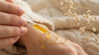 Woman applying golden serum oil to hand with dropper for skincare routine and moisturizing treatment on neutral background with dried flowers.