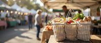 Wire shopping basket filled with fresh organic vegetables and groceries at outdoor farmers market with blurred shoppers and vendor stalls in background.