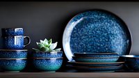Modern ceramic dinnerware set in deep blue and turquoise gradient with speckled glaze finish, stacked bowls, plates and cup with decorative succulent plant.