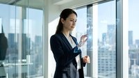 Professional Asian businesswoman in dark suit using smartphone in modern office with city skyline view through floor-to-ceiling windows.
