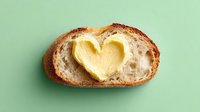 Fresh artisan bread slice with creamy butter shaped into heart on mint green background for food photography and culinary design projects.