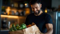 Young bearded man in black t-shirt smiling while holding paper bag with fresh groceries and vegetables in modern kitchen with warm ambient lighting.