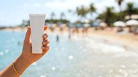 Close-up of hand holding blank white sunscreen tube at tropical beach with ocean and sandy shore in background for skincare product mockup.