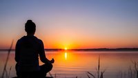 Woman meditating in lotus pose by calm lake at sunrise with golden sky reflection creating peaceful mindfulness moment for wellness and spiritual practice.