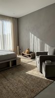 Minimalist bedroom interior with warm sunlight, neutral tones, jute rug, gray armchair, and a ginger cat resting on a modern pet bed.