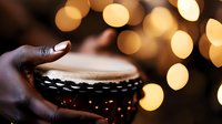 African American hands playing traditional djembe drum against warm bokeh lights background, creating atmospheric musical moment in soft golden glow.