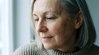 Mature woman with gray hair and freckles wearing a neutral sweater, looking thoughtfully to the side near a window for senior lifestyle, wellness, and portrait projects.