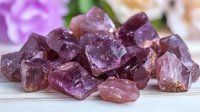 Raw amethyst crystal stones in purple and pink hues arranged on wooden surface with soft floral background, showcasing natural mineral texture and transparency.