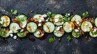 Fresh cucumber slices garnished with feta cheese crumbles, herbs, and balsamic glaze arranged in artistic pattern on dark rustic background.