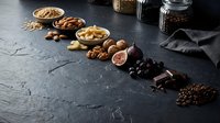 Assorted nuts, dried fruits and chocolate arranged on dark slate surface for healthy snacking and cooking ingredients display.