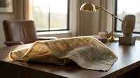 Cozy knitted blanket draped on wooden desk in warm sunlit home office with leather chair and brass lamp creating comfortable workspace atmosphere.