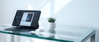 Modern glass desk workspace with open laptop displaying web design interface, small succulent plant and white coffee cup in minimalist office setting.