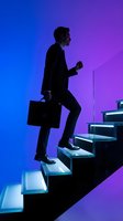 Silhouetted male professional in business suit climbing illuminated stairs while holding briefcase against vibrant blue to purple gradient background, representing career advancement and business growth.