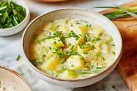 Creamy potato soup garnished with fresh chives and black pepper in ceramic bowl, served with bread on rustic wooden board against white background.