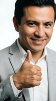 Middle-aged man with dark hair and olive complexion wearing gray blazer and white shirt giving thumbs up gesture against white background for business and corporate use.