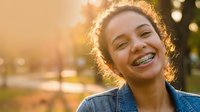 Happy mixed-race teenage girl with dental braces smiling outdoors in golden sunlight wearing denim jacket during sunset hour with warm bokeh background.