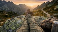 Hiker resting on mountain peak at sunset with boots extended, enjoying panoramic alpine valley views during outdoor adventure trek through rugged terrain.