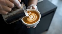 Barista creating latte art heart pattern in coffee cup with steamed milk from metal pitcher in cafe setting for coffee shop business and hospitality industry.