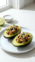 Halved avocados topped with shredded chicken, black beans, red onion and cilantro served on white plate with lime and sour cream.