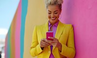 Young African American businesswoman in bright yellow suit using smartphone against colorful wall background, smiling at good news.