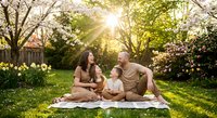 Happy mixed-race family with toddler enjoying spring picnic in blooming garden with cherry blossoms and daffodils during golden hour sunset.