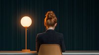 Young businesswoman sitting alone in dark room with glowing spherical lamp, back view, minimalist composition against green curtain background.