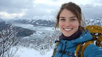 Young woman with brown hair and fair complexion, wearing blue winter jacket and mustard backpack, smiling at camera with snowy mountain landscape and frozen lake in background. Winter hiking and outdoor adventure portrait.