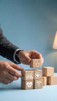 Hand placing wooden block with stethoscope icon onto stack of healthcare symbols including heart, house, medical cross and heartbeat against blue background for insurance and wellness concepts.