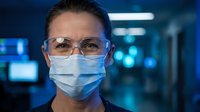 Professional healthcare worker wearing protective mask and safety glasses in modern medical facility with blue lighting and advanced equipment background.