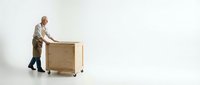 Senior warehouse worker pushing wooden crate on wheels across white floor. Professional logistics and shipping operations in clean industrial setting.