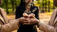 Diverse group of friends creating soap bubbles together in sunny park, hands reaching out to catch floating bubbles, joyful outdoor activity and friendship.