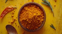 Ground turmeric powder in wooden bowl with dried red chili and fresh rosemary on yellow background, traditional Indian spice and seasoning.