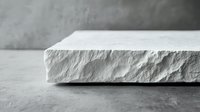 Minimalist white stone slab with rough textured edge on concrete background, dramatic side lighting creates subtle shadows emphasizing natural surface detail.