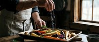 Professional chef seasoning roasted vegetables on wooden cutting board in rustic kitchen. Culinary preparation with fresh organic produce for healthy cooking.