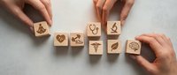 Hands arranging wooden blocks with medical healthcare symbols including stethoscope, heart, caduceus and brain icons on white background.